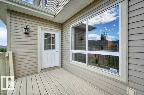 Outdoor deck featuring horizontal siding, a white entry door with glass panes, white-framed windows, and an exterior wall lantern - 14 4367 Veterans Way, Edmonton, AB 