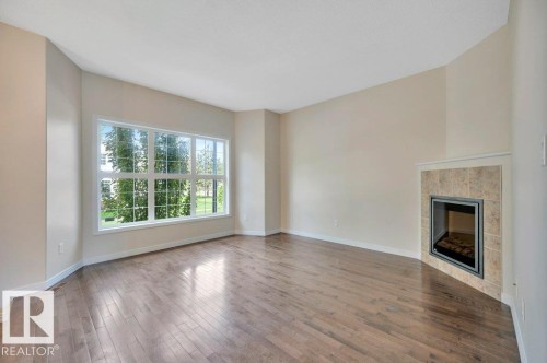 Spacious room featuring wood-finish flooring, a large multi-pane window, and a corner fireplace with a tiled surround - 14 4367 Veterans Way, Edmonton, AB 