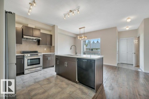Open-concept kitchen featuring dark wood-finish cabinetry, stainless steel appliances, a center island with sink and integrated dishwasher, and tile flooring - 14 4367 Veterans Way, Edmonton, AB 