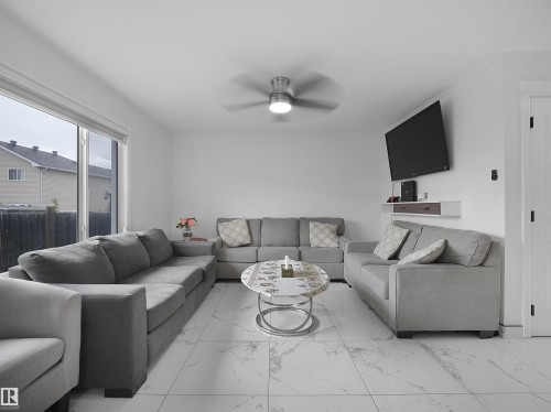 Spacious living area featuring large format white tile flooring, a contemporary ceiling fan with integrated lighting, and a prominent sliding glass door - 2418 32B Street, Edmonton, AB 