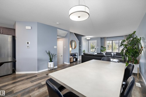 Open-concept living space featuring wood-finish flooring, a contemporary drum pendant light fixture, stainless steel refrigerator, and ample natural light from large windows - 2833 Chokecherry Place, Edmonton, AB 