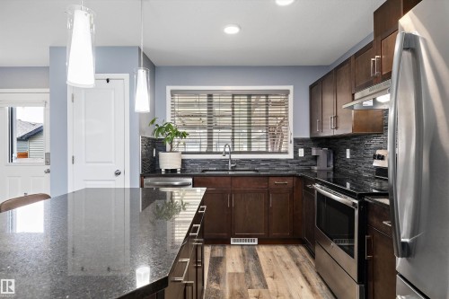 Kitchen featuring dark wood cabinetry, stainless steel appliances, a dark stone countertop island, wood-finish flooring, and a textured tile backsplash - 2833 Chokecherry Place, Edmonton, AB 