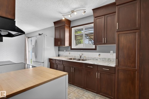 Kitchen featuring dark wood-finish cabinetry, light-toned countertops, a stainless steel double-basin sink, and a white tiled backsplash - 13427 114 Street, Edmonton, AB 