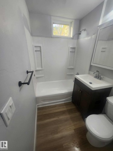 Bathroom featuring wood-finish flooring, a white bathtub with shower surround, a dark wood-finish vanity with an integrated sink, a wall-mounted mirror, and a window - 11308 94 Street, Edmonton, AB 