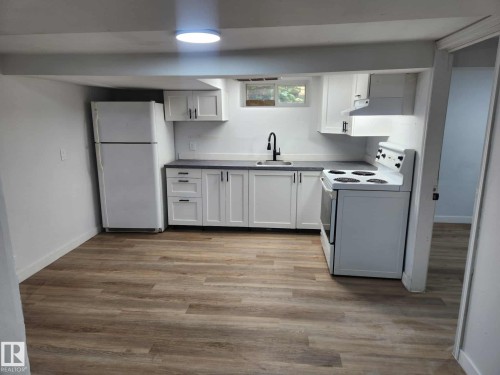 Kitchen featuring wood-finish flooring, white cabinetry with dark hardware, a matte black faucet, a window, and a white appliance suite - 11308 94 Street, Edmonton, AB 
