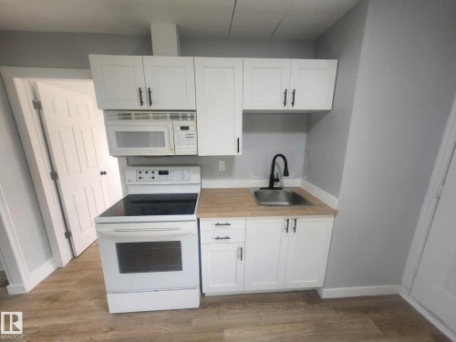 Efficient kitchen space featuring white shaker cabinetry, wood-finish countertops, stainless steel sink with a matte black faucet, a white electric range, and an over-the-range microwave - 11308 94 Street, Edmonton, AB 