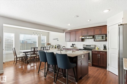 Open-concept kitchen and dining area featuring rich wood-finish flooring, a kitchen island with seating, stainless steel appliances, and ample natural light from multiple windows - 3685 Westcliff Way, Edmonton, AB 