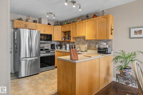 Kitchen featuring wood-finish cabinetry, stainless steel appliances, a center island with countertop seating, and tile flooring - 21 1850 Mill Woods Road E, Edmonton, AB 