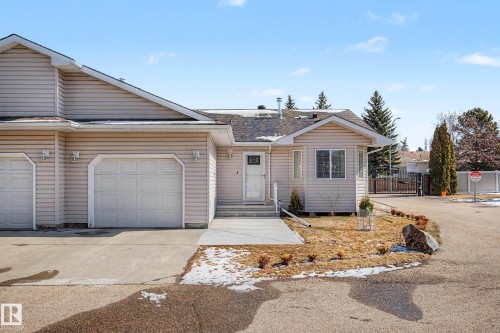 Residence featuring light beige siding, an attached single-car garage, a paved driveway, and a concrete walkway leading to the entry - 21 1850 Mill Woods Road E, Edmonton, AB 