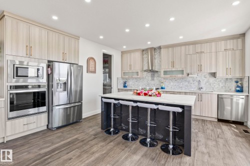 Kitchen featuring a large central island with a light-toned countertop, a full suite of stainless steel appliances, light wood-finish cabinetry, a patterned backsplash, and wood-finish flooring - 23 Elwyck Gate, Spruce Grove, AB 