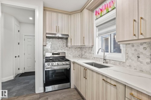 Modern kitchen featuring light wood-finish cabinetry, a stainless steel range with overhead ventilation, a hexagonal tile backsplash, and light-toned countertops - 23 Elwyck Gate, Spruce Grove, AB 