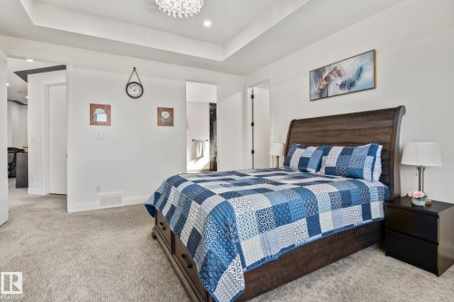 Carpeted bedroom featuring a tray ceiling with a chandelier, white walls, and dark wood-finish flooring visible through the doorway - 23 Elwyck Gate, Spruce Grove, AB 