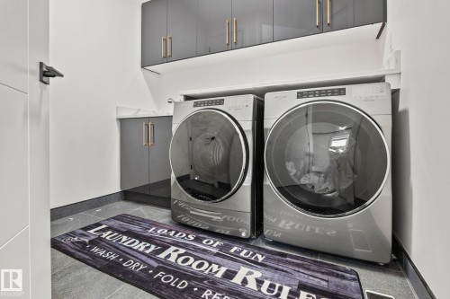 Laundry area featuring front-loading machines, built-in upper cabinetry with gold-tone hardware, a white countertop, lower gray cabinetry, and dark gray tile flooring - 23 Elwyck Gate, Spruce Grove, AB 