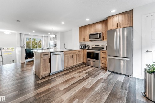 Open-concept kitchen featuring wood-finish cabinetry, stainless steel appliances, a center island with a built-in dishwasher, and wood-finish flooring - 116 3305 Orchards Link Sw, Edmonton, AB 