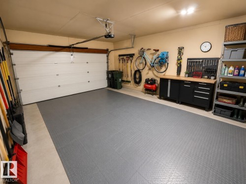 Spacious garage featuring a white overhead door, durable gray interlocking floor tiles, built-in workbench with storage, and integrated shelving units - 1 Munro Court, Leduc, AB 