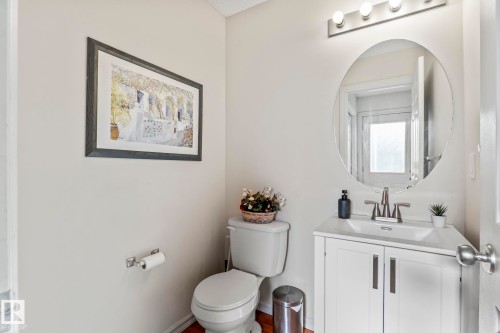 Bathroom featuring a white vanity with integrated sink, coordinating white toilet, brushed nickel fixtures, and a circular wall mirror with overhead bar lighting - 220 85St, Edmonton, AB 