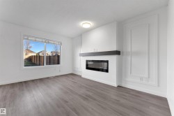 Spacious room featuring wood-finish flooring, a contemporary fireplace with a dark mantel, a large window, white walls, and decorative wall paneling - 