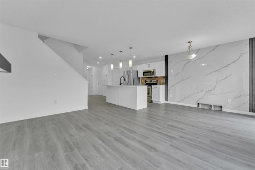 Open-concept living area featuring light wood-finish flooring, a contemporary kitchen with stainless steel appliances, a center island with pendant lighting, and a striking accent wall with a marble-effect finish - 5405 Crabapple Loop, Edmonton, AB 