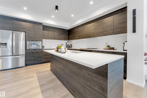Contemporary kitchen featuring a large center island with a light-toned countertop, wood-finish flooring, full-height dark wood-look cabinetry, a subway tile backsplash, and stainless steel appliances - 6203 47 Avenue, Beaumont, AB 