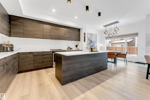 Open-concept kitchen featuring an expansive center island with countertop seating, wood-finish cabinetry, stainless steel appliances, light wood-finish flooring, and a modern rectangular chandelier - 6203 47 Avenue, Beaumont, AB 