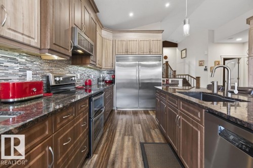 Gourmet kitchen featuring wood-finish flooring, two-tone cabinetry, stainless steel appliances including a built-in refrigerator and double oven, a tile backsplash, and dark stone countertops - 71 Ratelle Circle, St. Albert, AB 