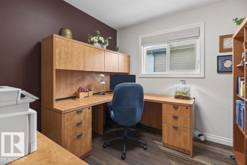 Dedicated workspace featuring a built-in wood-finish desk with integrated cabinetry, dark wood-finish flooring, a white trim window, and neutral wall tones - 71 Ratelle Circle, St. Albert, AB 