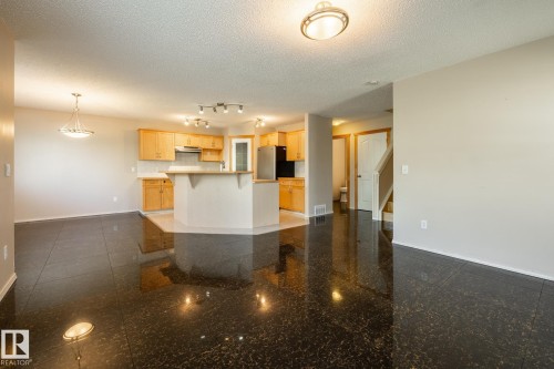 Spacious open-concept area featuring dark speckled flooring, a kitchen with light wood cabinetry, white countertops, and a central island with an extended breakfast bar - 55 Lamplight Cove, Spruce Grove, AB - Indoor Photo Showing Kitchen
