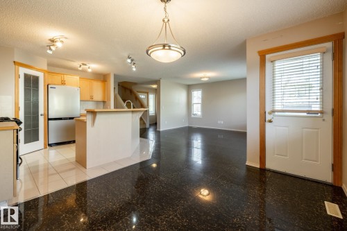 Bright interior featuring dark polished flooring, a kitchen with light wood-finish cabinetry, a built-in island with a sink, and a white entry door with privacy blinds - 55 Lamplight Cove, Spruce Grove, AB - Indoor Photo Showing Kitchen