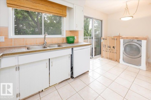 Kitchen area featuring light-toned square floor tiling, white cabinetry, a double basin stainless steel sink, and a large window with a woven shade - 4107 147 Street, Edmonton, AB 