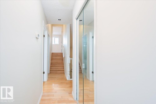 Well-lit hallway featuring wood-finish flooring, a full-length mirrored closet, and a staircase with painted white balusters leading to an upper level - 4107 147 Street, Edmonton, AB 
