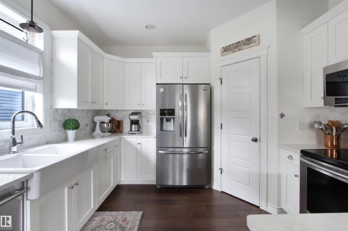 Modern kitchen featuring white shaker cabinetry, stainless steel appliances, a white farmhouse sink, light-toned countertops, and dark wood-finish flooring - 34 Falcon Road, Cold Lake, AB 