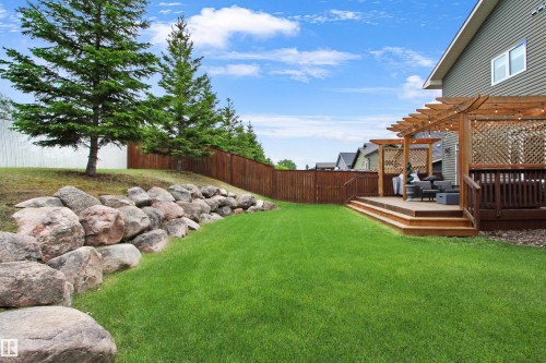 Expansive backyard featuring a multi-level wood deck with a pergola, a rock retaining wall, mature evergreen trees, and a privacy fence - 34 Falcon Road, Cold Lake, AB 