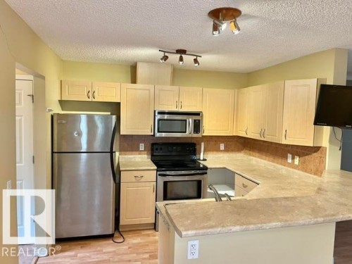 U-shaped kitchen featuring light wood cabinetry, stone-look countertops, stainless steel appliances, and a tiled backsplash - 1 5281 Terwillegar Boulevard, Edmonton, AB 