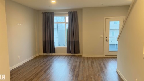 Spacious room featuring wood-finish flooring, light neutral wall tones, a large window with privacy coverings, and a glass-paneled exterior door - 90 804 Welsh Drive, Edmonton, AB 