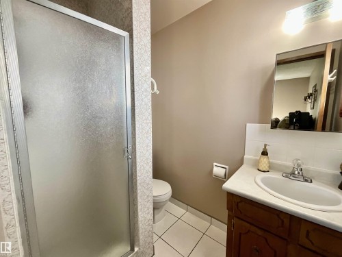 Bathroom featuring a shower with a frosted glass door, a wood-finish vanity with a white countertop and integrated sink, a wall-mounted mirror, and white tile flooring - 11031 166 Avenue, Edmonton, AB 
