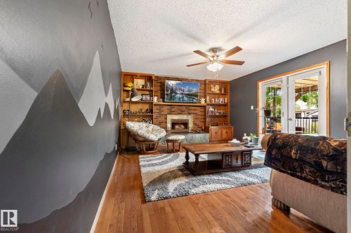Living area featuring wood-finish flooring, a brick-surround fireplace, built-in shelving, a ceiling fan, and double French doors opening to an exterior space - 9940 83 St, Fort Saskatchewan, AB 