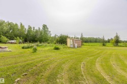 Expansive green space featuring a utility shed with a wooden facade, a well housing, a fenced perimeter, and mature evergreen trees - 