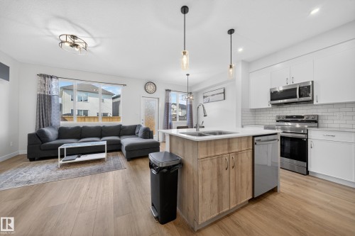 Open-concept living and kitchen area featuring wood-finish flooring, white cabinetry, stainless steel appliances, and a kitchen island with a double basin sink - 1684 12 Street, Edmonton, AB 