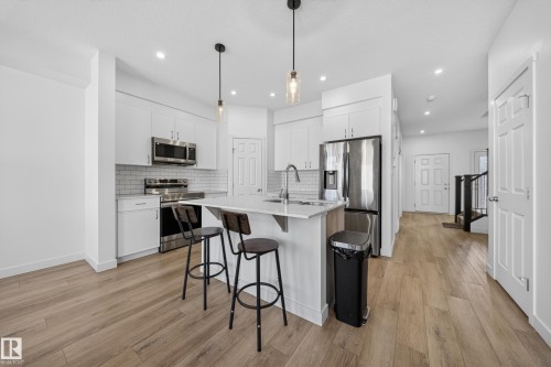 Modern kitchen featuring a central island with seating, stainless steel appliances, white cabinetry, subway tile backsplash, and wood-finish flooring - 1684 12 Street, Edmonton, AB 