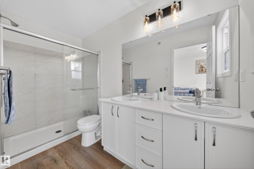 Contemporary bathroom featuring a double vanity with white cabinetry and integrated sinks, a wide mirror, a three-light fixture, a toilet, and a shower-over-tub with sliding glass doors and white tiled surround - 1684 12 Street, Edmonton, AB 