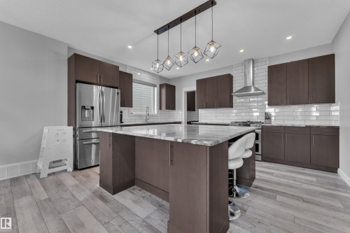 Kitchen featuring a large island with stone countertop, stainless steel appliances, dark wood-finish cabinetry, white subway tile backsplash, and light wood-finish flooring - 17551 64 Street, Edmonton, AB 