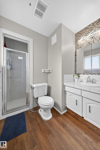 Full bathroom featuring a glass-enclosed shower, white toilet, wood-finish flooring, a white vanity with an integrated sink, and a decorative tiled backsplash - 2832 35 Street, Edmonton, AB 