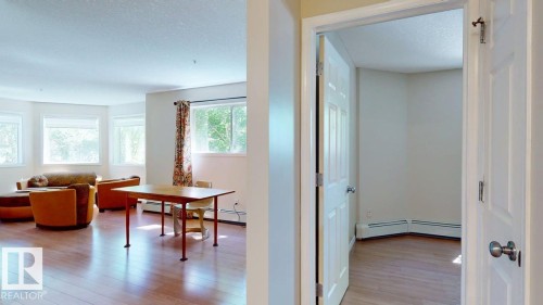 Bright living area featuring wood-finish flooring, multiple windows, and a doorway leading to a separate room with baseboard heating - 201 11408 108 Avenue, Edmonton, AB 
