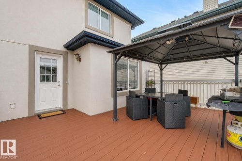 Spacious wood-finish deck featuring a permanent gazebo structure, a white exterior door with glass panes, an exterior wall sconce, and a sliding glass window - 710 Glenwright Court, Edmonton, AB 