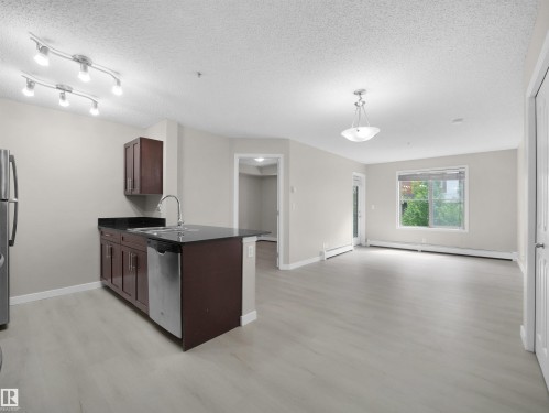 Open-concept living space featuring light wood-finish flooring, a kitchen island with dark cabinetry and countertop, stainless steel appliances, and a large window providing natural light - 220 12035 22 Avenue, Edmonton, AB 