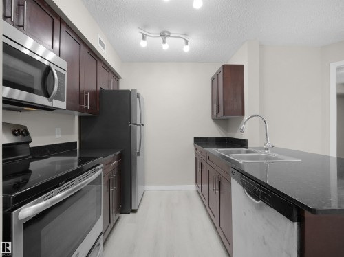 Galley kitchen featuring dark wood cabinetry, stainless steel appliances, black countertops, and a double basin sink - 220 12035 22 Avenue, Edmonton, AB 