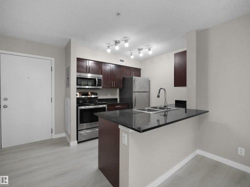 Kitchen featuring dark wood cabinetry, stainless steel appliances, and a black countertop with an undermount double sink - 220 12035 22 Avenue, Edmonton, AB 