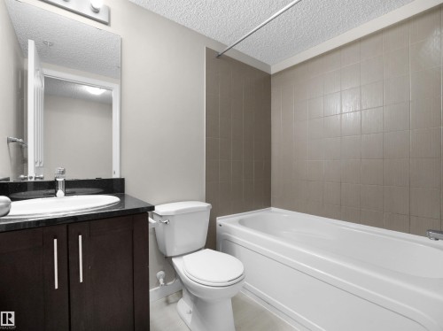 Bathroom featuring a white bathtub, tiled shower surround, dark wood-finish vanity with a black countertop, undermount sink, and a wall-mounted mirror - 220 12035 22 Avenue, Edmonton, AB 