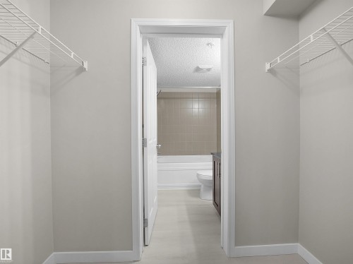 Walk-through closet with built-in wire shelving, neutral wall paint, and white trim, opening to a full bathroom featuring a white bathtub, tiled shower surround, and a dark wood-finish vanity - 220 12035 22 Avenue, Edmonton, AB 