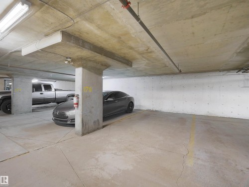 Spacious concrete parking area with designated parking spaces, visible fire extinguisher, and overhead fluorescent lighting - 220 12035 22 Avenue, Edmonton, AB 